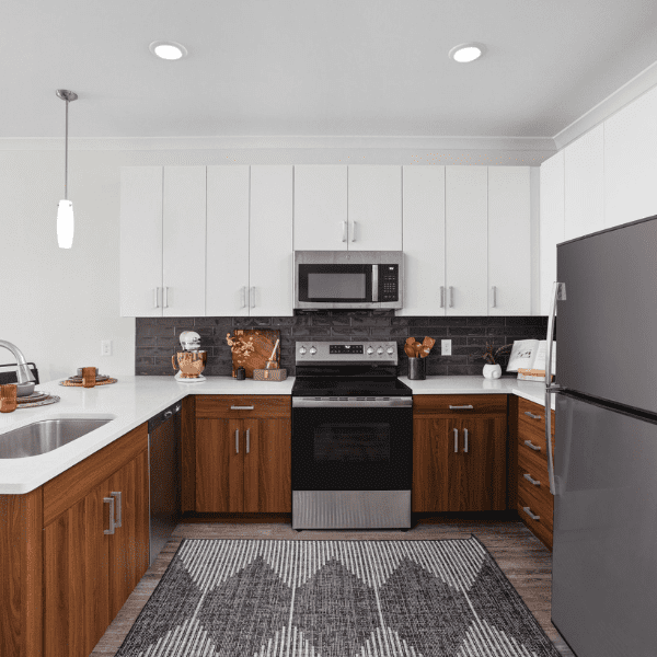 A modern living room with a loft design features a metal staircase leading to an upper level. Two large windows with blinds allow natural light to fill the space. The room includes a gray couch, a coffee table with a glass fireplace, and a floor lamp.